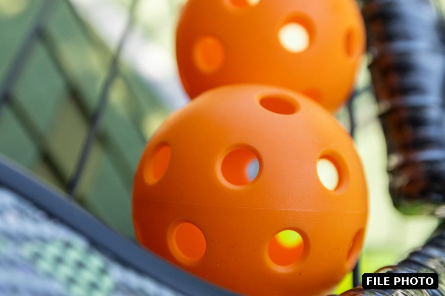 Two orange pickleballs resting on a pickleball paddle by a court fence