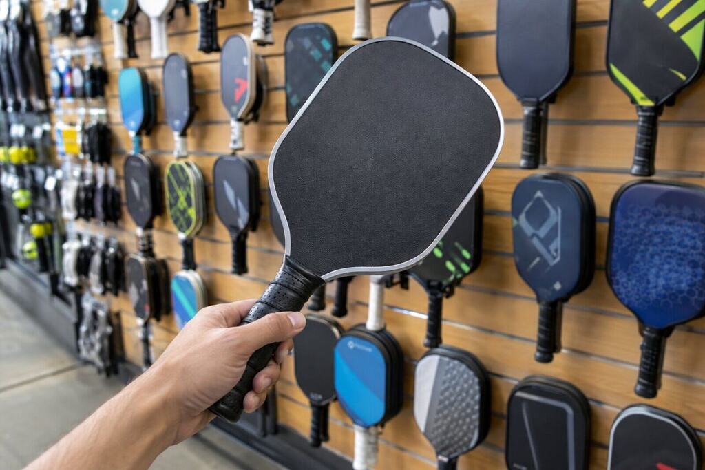 A hand holding a pickleball paddle over a display of different paddle shapes and thicknesses at a shop