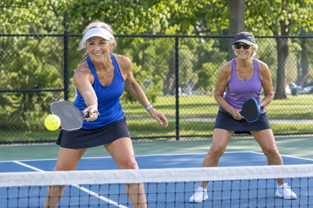 Two active older women playing pickleball doubles on an outdoor court, smiling mid-rally