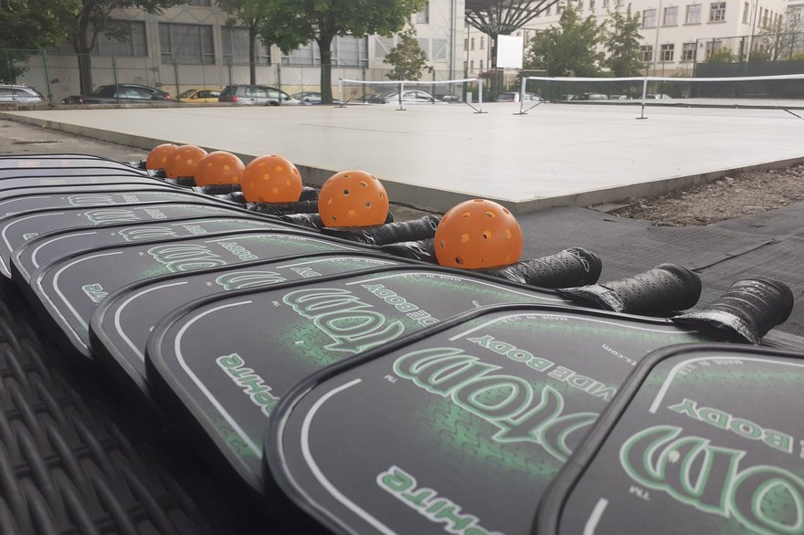A row of pickleball paddles and orange pickleballs lined up beside an outdoor court